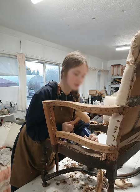 Stagiaire dégarnissant la carcasse d’un fauteuil ancien au marteau dans l’atelier de tapisserie